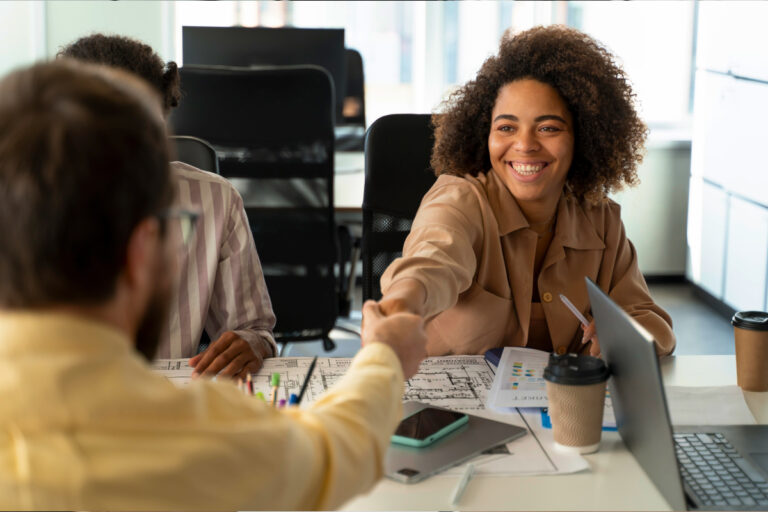 Imagem de uma mulher e um homem se cumprimentando enquanto sorriem, ilustrando o RH fazendo a gestão de benefícios da empresa, no escritório corporativo.