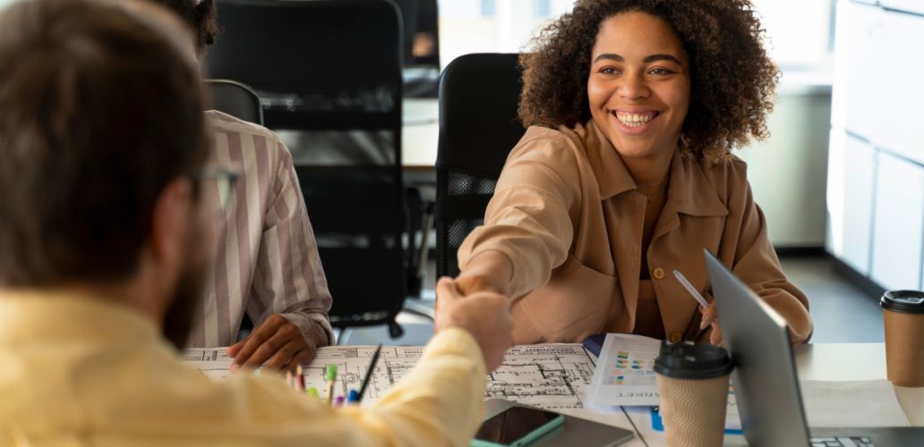 Imagem de uma mulher e um homem se cumprimentando enquanto sorriem, ilustrando o RH fazendo a gestão de benefícios da empresa, no escritório corporativo.